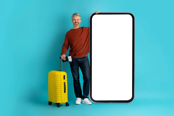 チェックインオンライン 白い空の画面で巨大なスマートフォンを使用してカジュアルな服装の旅行者で幸せな中年の男性 黄色の荷物を運ぶ チケットでパスポートを保持 モックアップ 青のスタジオの背景 — ストック写真