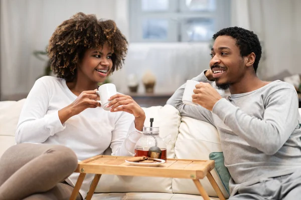 幸せな美しい若い黒のカップルが自宅でソファに座って お茶を飲んで会話をしてください パジャマでアフリカ系アメリカ人の千年の男と女を愛しますドリンクを楽しみます リビングルームのインテリア — ストック写真