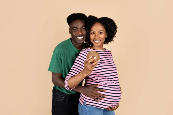 笑千年的非洲裔美国丈夫抱着大肚子的女人 女士抱着椰子 被米色背景隔离 工作室 广告和服务 — 图库照片