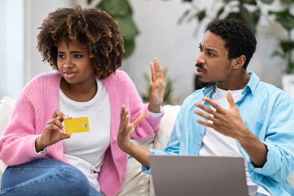 stock image Angry handsome young black man husband with laptop computer on his lap scolding his wife shopaholic, irritated african american woman holding bank card and showing boyfriend stop gesture