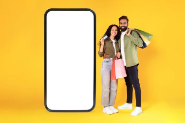 Satış reklamı. Alışveriş poşetleri ve kredi kartı ile mutlu Arap Alıcılar çift boş ekranlı dev akıllı telefonun yanında sarılıyor, sarı stüdyo arka planında kamera pozuna gülümsüyor. Kolaj