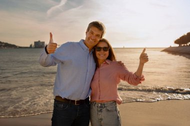 Güneşin batışında kumsalda poz veren mutlu olgun çift ve kamerada başparmaklarını yukarı kaldıran neşeli orta yaşlı eşler tatil beldesi tavsiye edilen seyahat acentesi, yer ayırtma,