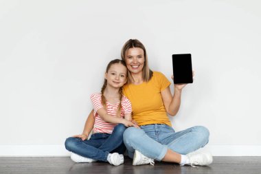 Çevrimiçi reklam. Mutlu Genç Anne ve Küçük Kız, evde yerde dinlenirken boş ekranlı boş dijital tablet tutuyorlar. Anne ve bayan çocuk fotokopi alanını gösteriyor.