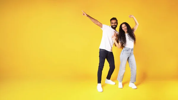 黄色のスタジオの背景にあるコピースペースで 陽気な中東の若いカップルがポーズをとって指を脇に向けています 楽しい千年の男と女の広告テキストの提供の完全な長さのショット パノラマ — ストック写真