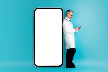 Sağlık Hizmetlerinde Teknoloji ve İletişim. Orta yaşlı doktor mavi arka planda beyaz ekranlı akıllı telefonla poz veriyor. Dijital tablet kullanıyor, iş arkadaşlarıyla, hastalarla, modellerle iletişim kuruyor.