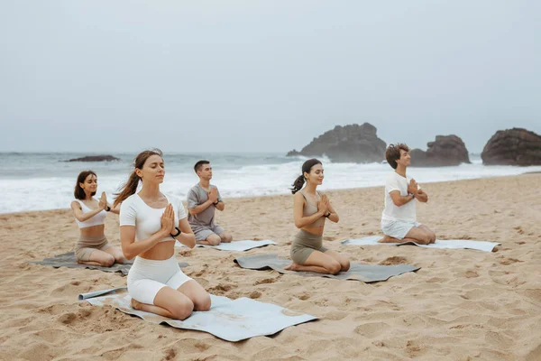 stock image Yoga group sitting and meditating on the beach, practicing asana with joined hands, people enjoying morning training at coastline. Concentration and serenity concept