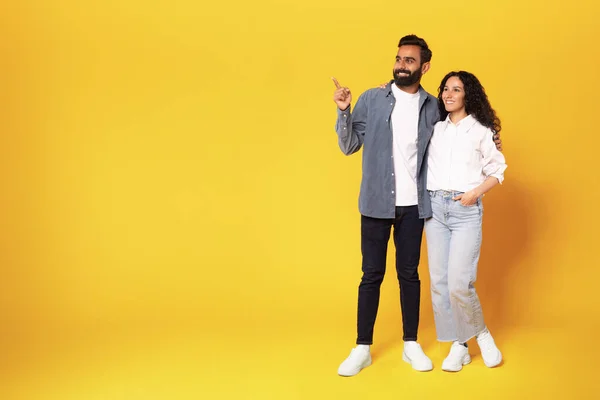 これを確認しろ 黄色のスタジオの背景に立つと抱擁のための無料のコピースペースを示す脇にカジュアルなポインティングフィンガーで幸せなアラビア語のカップル 全長撮影 広告バナー — ストック写真