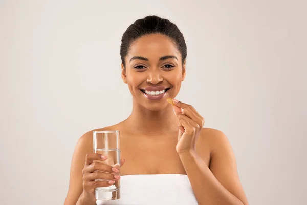 幸せな美しい若いアフリカ系アメリカ人女性の美しい肖像画バスタオルに包まれた灰色の背景に孤立立って 美しさを示す錠剤カプセル サプリメント 水のガラスを保持 — ストック写真