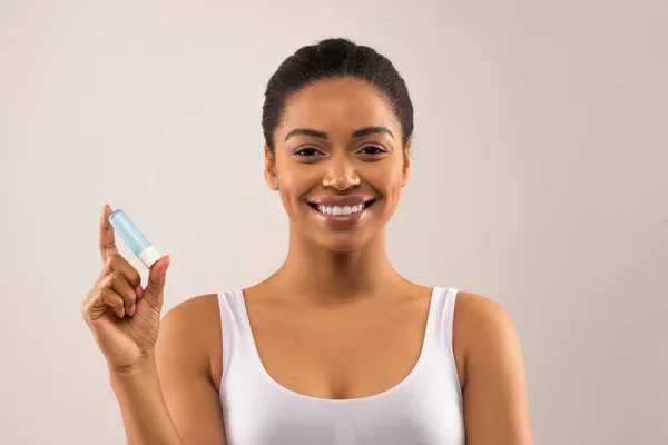 明るく美しい若いアフリカ系アメリカ人女性を身に着けている白いトップ保持口紅と笑顔 黒の女性適用衛生リップバーム上の光の背景 コピースペース — ストック写真