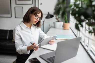 Konsantre genç bayan çalışan çalışma masasında oturuyor, dizüstü bilgisayar ve dijital ped kullanıyor, rahat ofis içinde çalışıyor, kopyalama alanında çalışıyor. İş dünyasındaki modern teknolojiler
