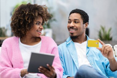 E-ticaret konsepti. Güzel gülümseyen genç siyah çift evlerinden internetten alışveriş yapıyorlar, rahat oturma odasındaki kanepede oturuyorlar, modern dijital tablet ve banka kartı kullanıyorlar, boşlukları kopyalıyorlar.