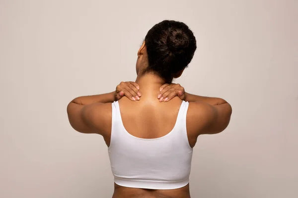 Neck pain muscle stress and strain, office syndrome concept. Stressed african american woman in white top massaging neck, back view, isolated on grey background