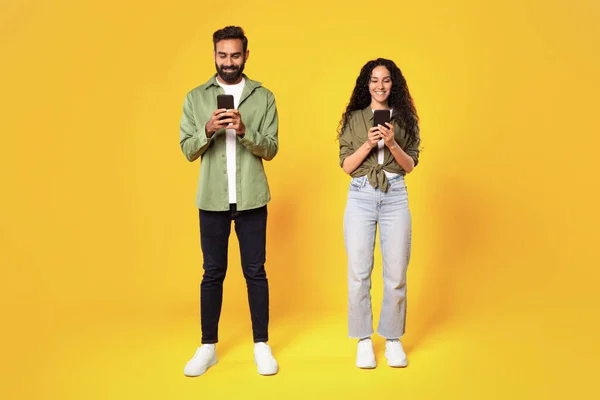 モバイル通信 黄色のスタジオの背景に立っているスマートフォンを保持している中東のカップルのテキストを微笑む 携帯電話でアプリケーションを使用してアラブの男と女 フルレングスショット — ストック写真
