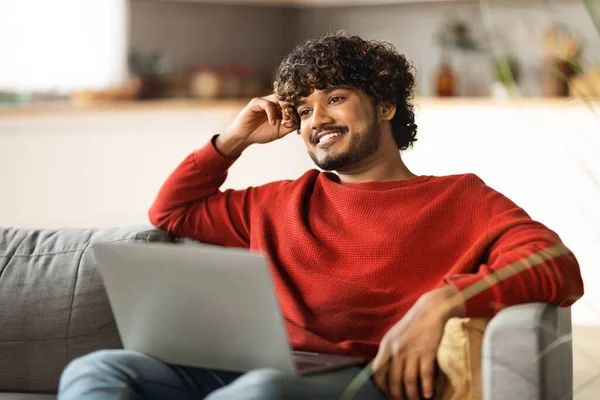 stock image Smiling Handsome Indian Guy Resting On Couch At Home With Laptop, Happy Young Eastern Male Blogger Thinking About New Post In Social Media, Using Computer For Online Work, Free Space