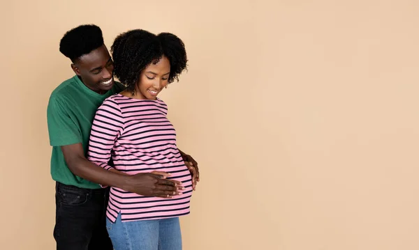 快乐的千年非洲裔美国丈夫抱着大肚子的女人 感觉婴儿 孤立在米色的背景下 工作室 不孕症治疗 — 图库照片