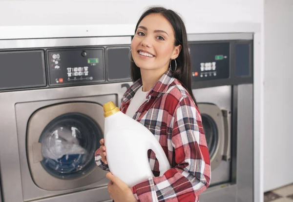 洗涤剂服务 快乐的年轻女士拿着洗涤剂瓶对着相机微笑 广告洗衣店 站在并放置在工业洗衣机的近旁 — 图库照片
