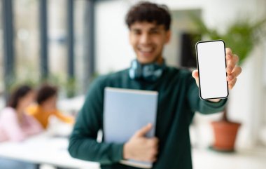 Eğitici reklam. Beyaz beyaz ekranlı akıllı telefon gösteren mutlu erkek öğrenci, model, seçici odaklanma. Arka planda sınıf arkadaşlarıyla birlikte çalışan bir adam.