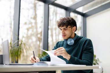 Avrupa 'lı genç adam ödevini defter içinde yazıyor, bilgisayardan internet üzerinden webinars izliyor, modern kütüphanede okuyor, boş alan arıyor.