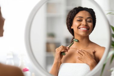 Güzelim siyahi kadın, yeşil taşlı yuvarlak boyunla masaj yapıyor, gülümseyen Afro-Amerikan kadın aynaya bakıyor ve cilt bakımı için güzellik aletini kullanıyor.