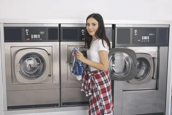 洗衣店概念 年轻的欧洲妇女站在Laudromat房间的洗衣机旁 手里拿着一件件衣服 看着相机的室内 夫人在洗衣房和干衣机旁 — 图库照片