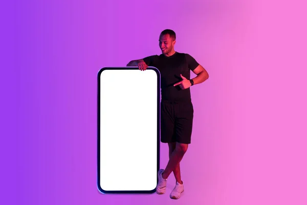 ワークアウトアプリケーション スポーティな黒の若い男は 紫のスタジオの背景の上に巨大なスマートフォンや空の画面を指してリース 携帯電話の近くのフィットネス広告モバイルアプリのスポーツマン モックアップ — ストック写真