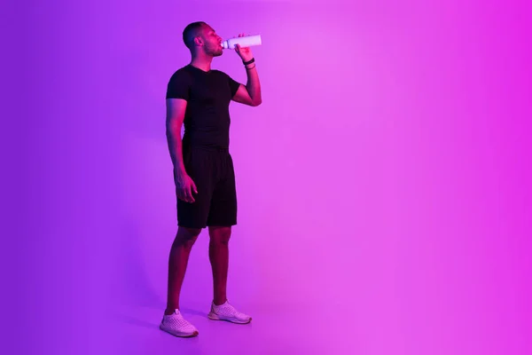 身穿运动服的年轻黑人运动员穿着紫色背景的运动服喝水 摆好姿势 站在空旷的空间附近 全身上下都是运动后保湿的运动健将 — 图库照片
