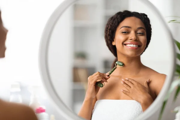 家でグリーンストーンジェイドローラーで美しい若い黒人女性マッサージネック 鏡を見て千年のアフリカ系アメリカ人女性を笑顔とスキンケアのための美容ツールを使用して — ストック写真