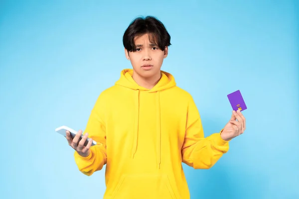 不満な買い手 スマートフォンとクレジットカードを持っている韓国の10代の男性は 失望と負の感情を表現し 青いスタジオの背景に銀行口座にお金を持っていません 失敗した取引 — ストック写真