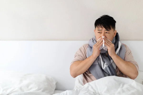 健康的中年男子 身穿睡衣 脖子上缠着围巾 躺在床上打喷嚏 患有感冒 流鼻涕 望着复印空间 — 图库照片