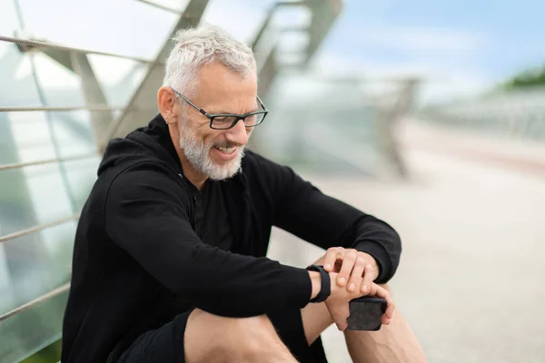 Senior man sitting on footbridge, checking smart watch, copy space. Stopwatch, positive elderly sportsman and runner check time, heart rate or monitor healthy body progress on data app