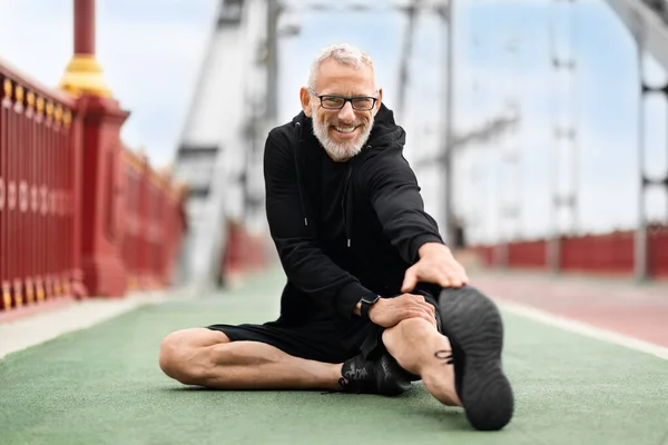 stock image Happy healthy sporty senior man wearing black sportswear sitting on ground at stadium, stretching body, reaching leg with hand while exercising outdoor, smiling at camera, copy space