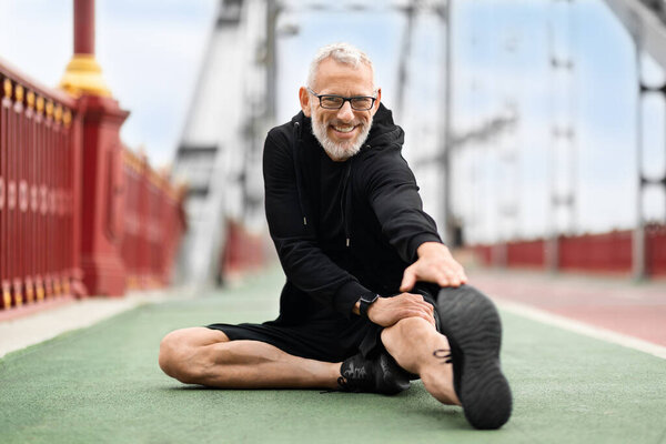 Happy healthy sporty senior man wearing black sportswear sitting on ground at stadium, stretching body, reaching leg with hand while exercising outdoor, smiling at camera, copy space