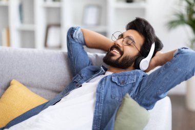 Yakışıklı sakallı Hintli adam evdeki kablosuz kulaklıklarla müzik dinliyor, arkası kapalı koltuğa yaslanmış gülümseyen genç doğu adamı, favori müzik listesinin keyfini çıkarıyor.