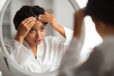 Endişeli genç siyah kadın saçlarını evde kontrol ediyor, kepek problemi var, Afro-Amerikan kadın aynanın yanında dikiliyor, Flaky Scalp çekiyor, yansımaya odaklanıyor.