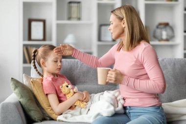 Mevsimsel grip. Endişeli bir anne, hasta bir kadına sıcak çay verip alnına dokunuyor, küçük kızını umursuyor, acı çekiyor, soğuk algınlığı geçiriyor ya da grip oluyor.