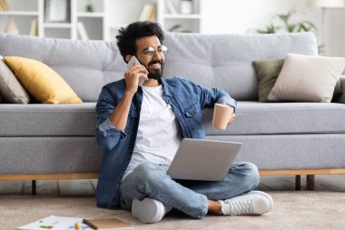 Uzaktan kumanda. Cep telefonuyla konuşan ve evde dizüstü bilgisayarla çalışan gülümseyen Hintli adam, telefon görüşmesi yaparken kahve içen, uzaktan çalışmanın keyfini çıkaran,
