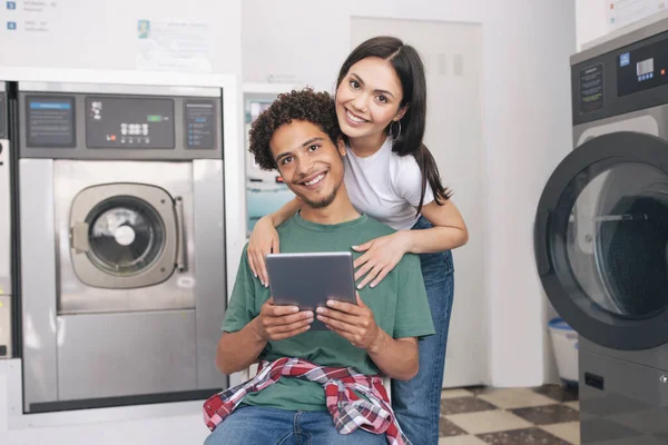 简单的洗衣 在公共洗衣房用洗衣机在洗衣房用平板电脑拥抱多种族的年轻家庭伴侣 在相机前微笑 — 图库照片