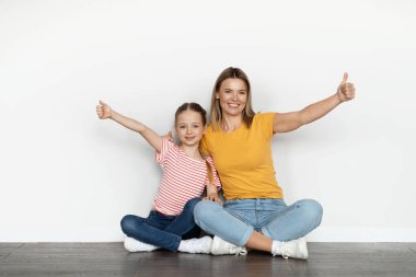 Mutlu, genç bir anne ve küçük bir kız kameranın önünde başparmaklarını kaldırırken ve yerde dinlenirken neşeli, beyaz bir anne ve tatlı bir kız çocuğu bir şey öneriyor.
