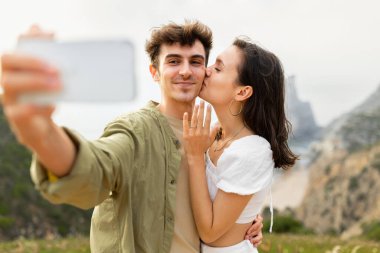 Nişanlandıktan sonra mutlu bir çift cep telefonuyla selfie çekiyor, yanağından öpen bir kadın, deniz kıyısındaki mükemmel teklifi yakalıyor, okyanus kıyısındaki kayalıklarda duruyor.