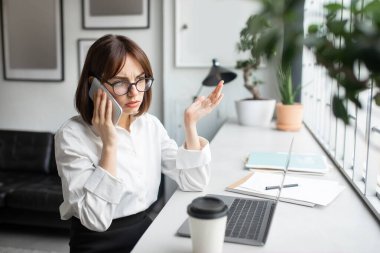Uzaktan çalışma stresi. Cep telefonuyla konuşan ve dizüstü bilgisayar kullanan endişeli iş kadını, müşteri desteğini arıyor, bilgisayar üzerinde çalışırken kadın serbest çalışanın yaşadığı sorunları vurguluyor