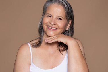 Beyaz saçlı, mutlu, yaşlı, beyaz bir kadın elleriyle yüzüne dokunuyor. Yaşlanma karşıtı muameleden hoşlanıyor. Kırışıklık kavgası, kahverengi arka plan, stüdyo. Güzellik, kaplıca, menopoz.