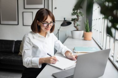 Gülümseyen iş kadını dizüstü bilgisayar kullanıyor ve modern ofisteki işyerinde notlar alıyor, iş sitelerini izleyen ve fotokopi defterine bir şeyler yazan bir kadın, masa başında oturuyor.