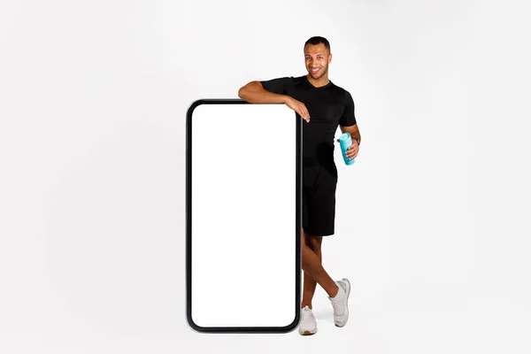 ワークアウトアプリケーション広告 黒フィットネス男は空の画面で大きな携帯電話でリース水のボトルを保持 スタジオで白い背景に立って フルレングスショット モックアップ — ストック写真