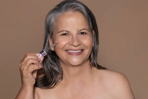 灰色の髪を持つ陽気な高齢者の白人女性はアンチエイジング治療を楽しんでいます血清を使用しています 茶色の背景に孤立 スタジオ 美容ケア しわの戦いとプロの化粧品 — ストック写真