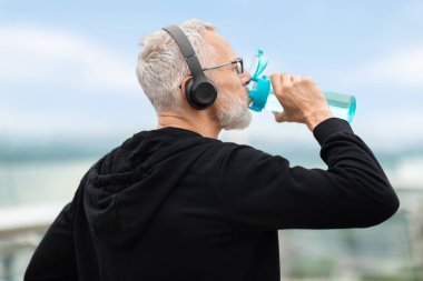 Spor yaparken, yakışıklı gri saçlı yaşlı sporcunun su içtiği yakın plan fotoğraf. Parkta koşan atletik son sınıf öğrencisi, siyah spor kıyafetleri, gözlükler, kablosuz kulaklıklar takıyor.