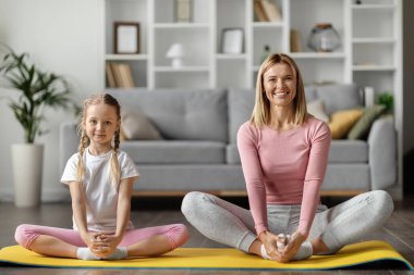 Güzel, genç bir anne ve sevimli küçük bir kız evde birlikte geriniyorlar mutlu bir aile annesi ve kız çocuğu kelebek egzersizi yapıyor ve kameraya gülümsüyor oturma odasında egzersiz yapıyor,