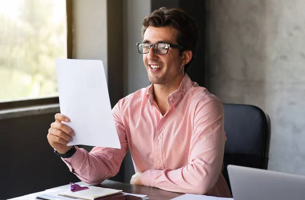職場でピンクのフォーマルシャツと眼鏡を身に着けている幸せなハンサムな千年紀のビジネスマン 紙を保持し オフィスでの対応を読み取り コピースペース 企業生活 — ストック写真
