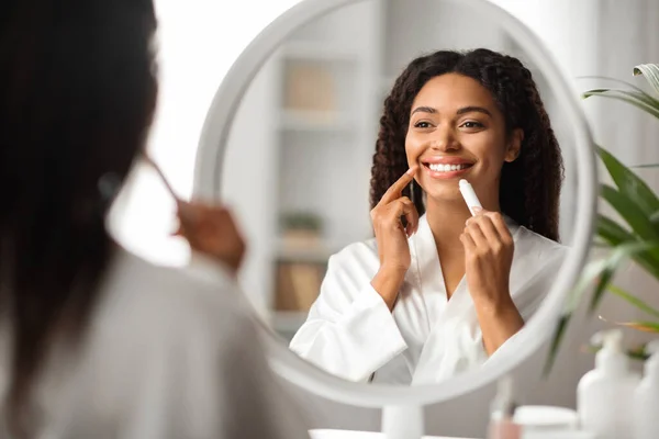 美しい黒の女性は鏡を見て 保湿リップバームを適用します 魅力的なアフリカ系アメリカ人女性がチャップスティックを保持笑顔 美容ルーチンを作りながら 衛生的なリップスティックを使用して — ストック写真