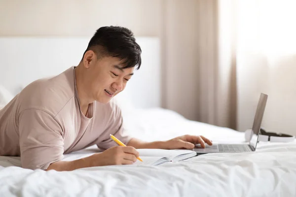 积极英俊的亚裔中年男子在家参加网络研讨会 躺在床上 使用笔记本电脑 在笔记本中记笔记 穿睡衣 复制空间 成人在线教育 — 图库照片