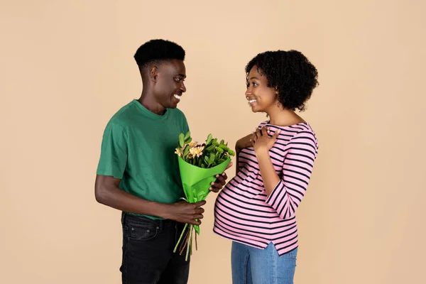 微笑的千年黑人把一束鲜花送给大肚子的女士 被米色背景 工作室隔离 家庭日期 庆祝周年 祝贺生日 期待孩子 — 图库照片
